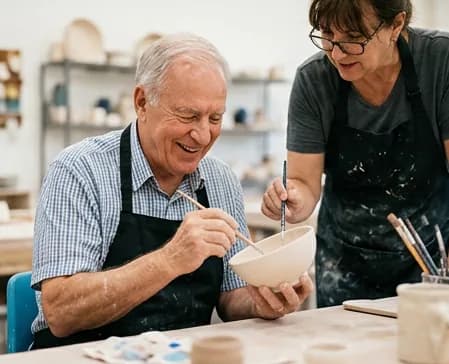 An older man painting a ceramic bowl while a workshop facilitator guides him
